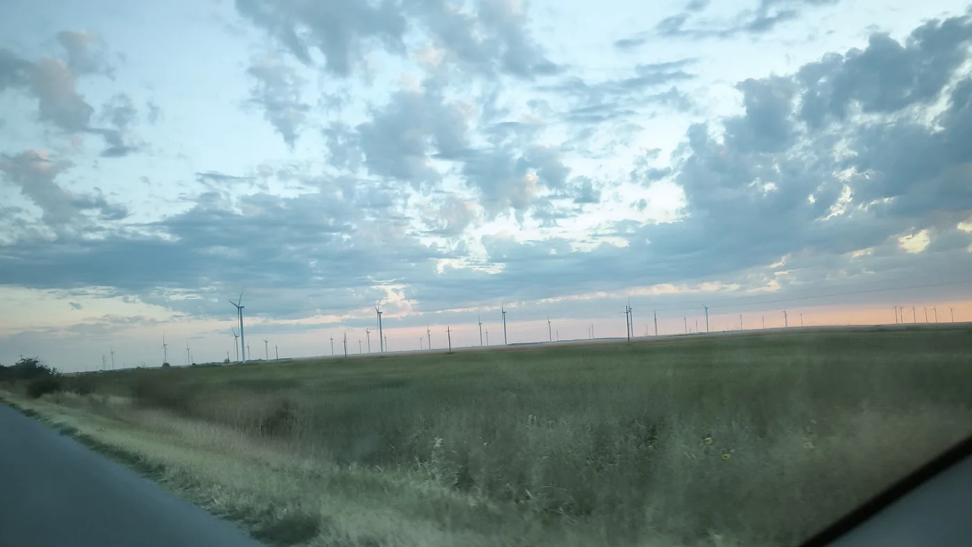 Flat grassland under a cloudy dawn sky with a long row of wind turbines on the horizon, seen from a moving car