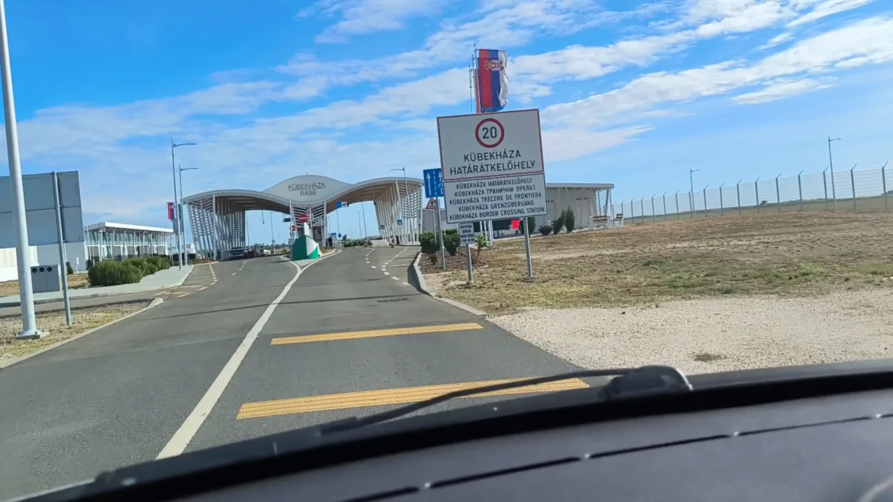 The Kubekhaza border crossing in daylight: a 20 km/h sign naming the crossing in several languages, the curved control canopy and a fence beside the road