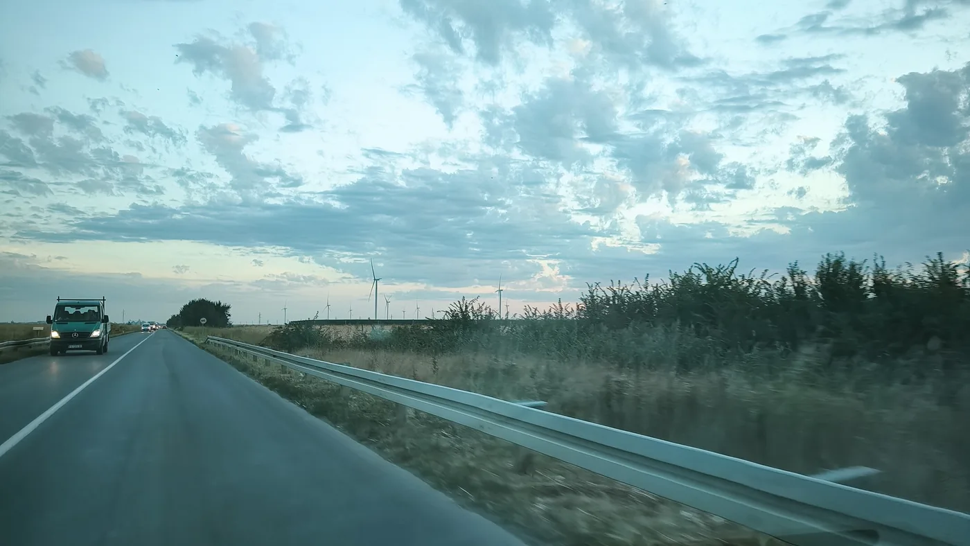 A two-lane country road at dawn in Serbia seen through a car windscreen, with a guardrail, a van approaching and wind turbines on the horizon