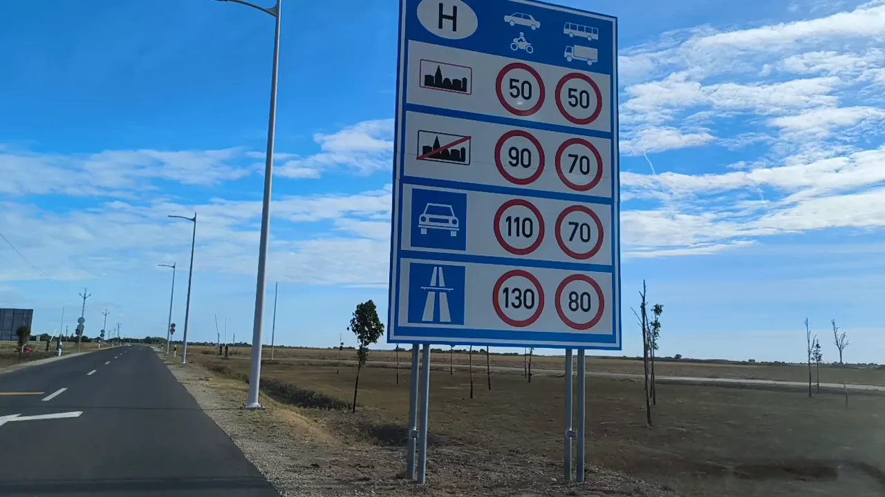 A blue Hungarian entry sign with speed limits beside a new, empty road across flat fields under a blue sky
