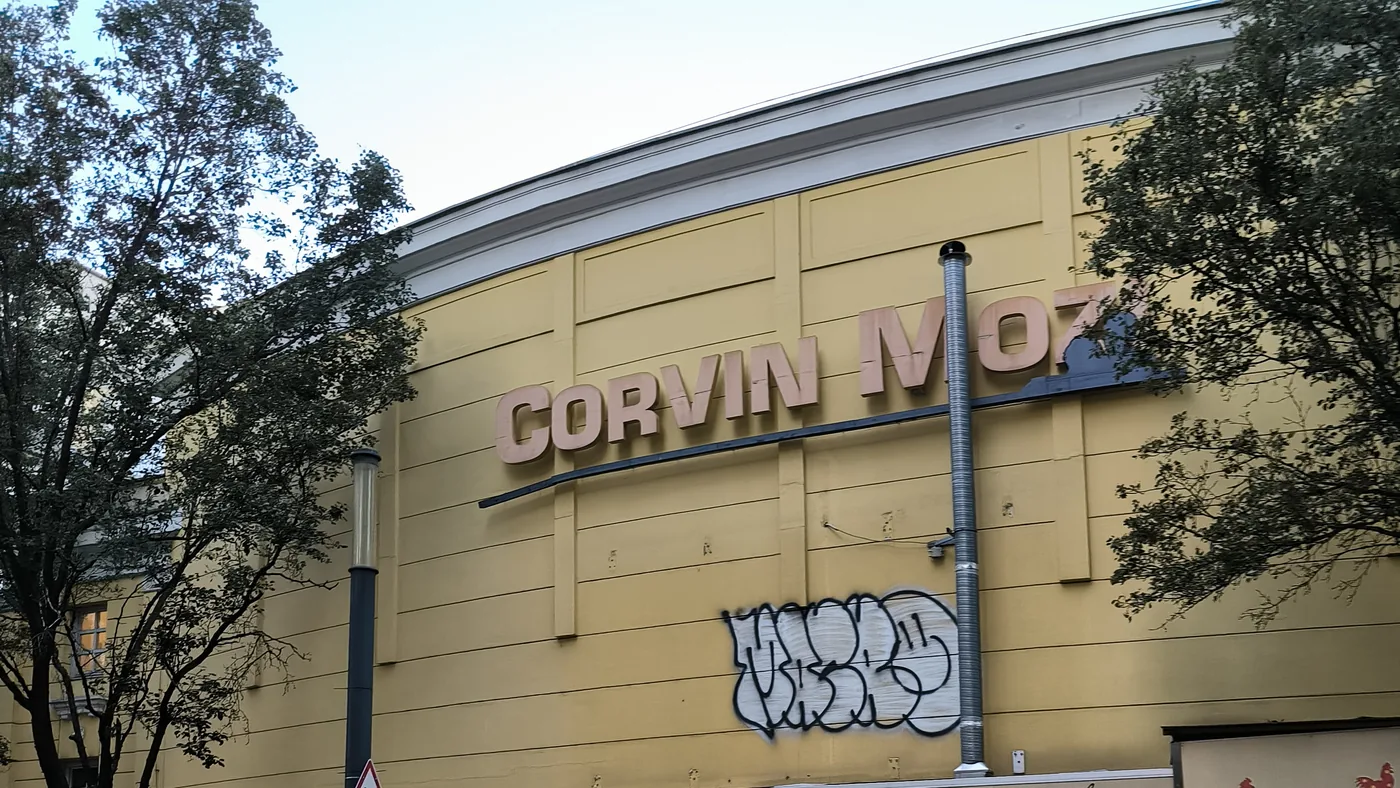 The yellow curved facade of the Corvin cinema in Budapest with its lettering, graffiti and a closed grilled-chicken kiosk on the square