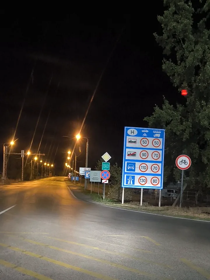 A blue Hungarian border entry sign at night showing speed limits of 50, 90, 110 and 130 km/h, beside a road lit by street lamps