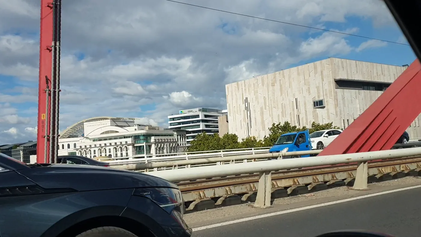 Pale stone and white modern buildings on the riverbank seen past a bridge railing, with a red mast in the foreground