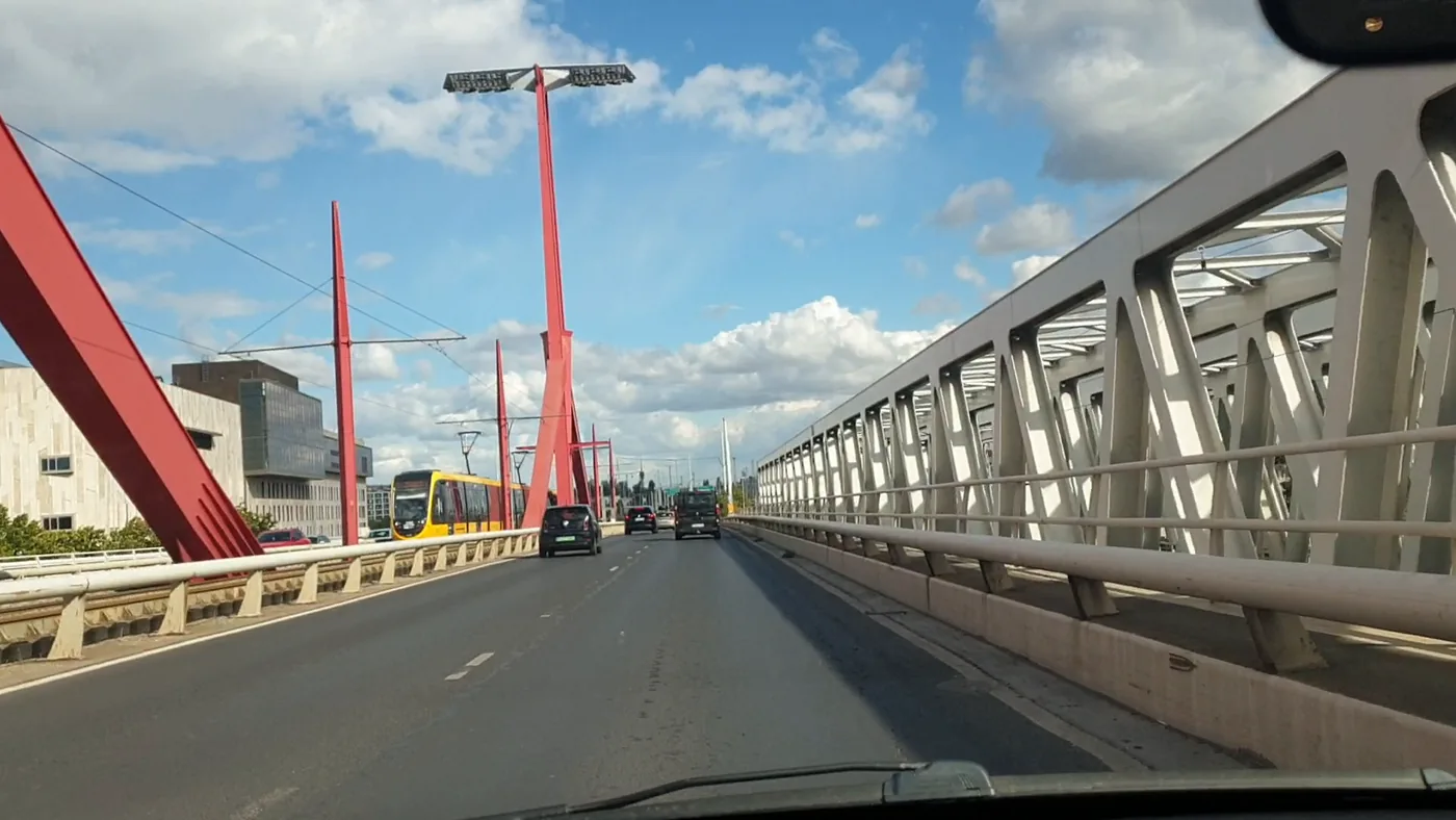 A steel truss bridge with tall red lamp masts, a yellow tram and light traffic ahead under a blue sky with clouds
