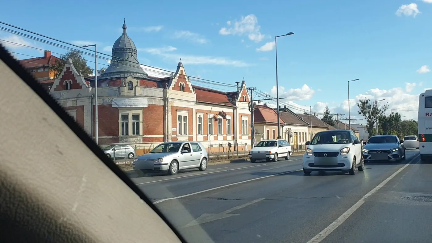 A red-brick building with a grey domed tower beside a main road, cars waiting at a junction under a blue sky