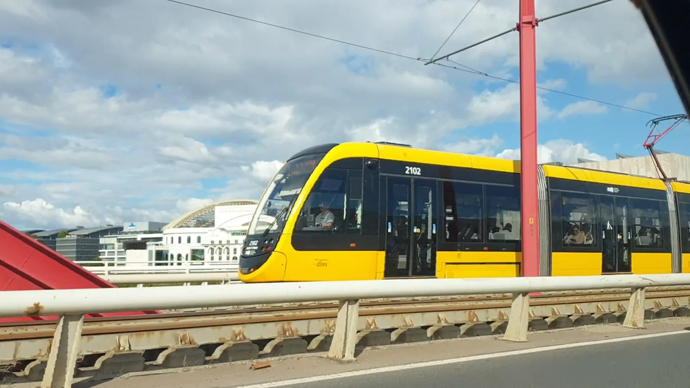 A yellow articulated Budapest tram, number 2102, beside the road on a bridge next to a red lamp mast