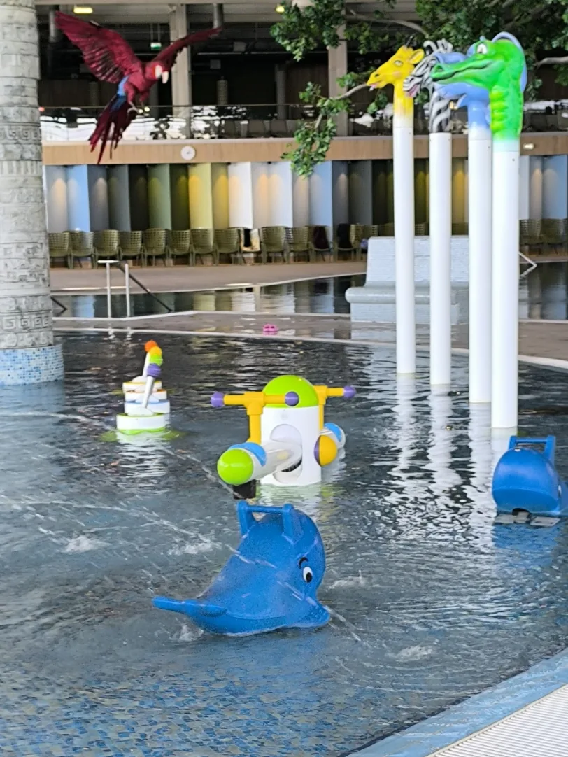 A shallow children's pool with water toys, a blue dolphin and animal-head fountains, and a model parrot hanging above