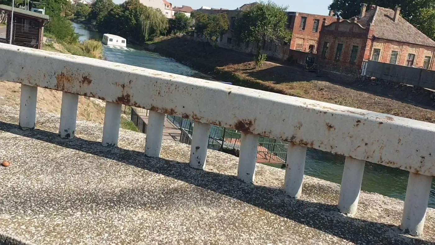 A rusted white bridge railing over a green canal with a small boat, old brick buildings on the far bank