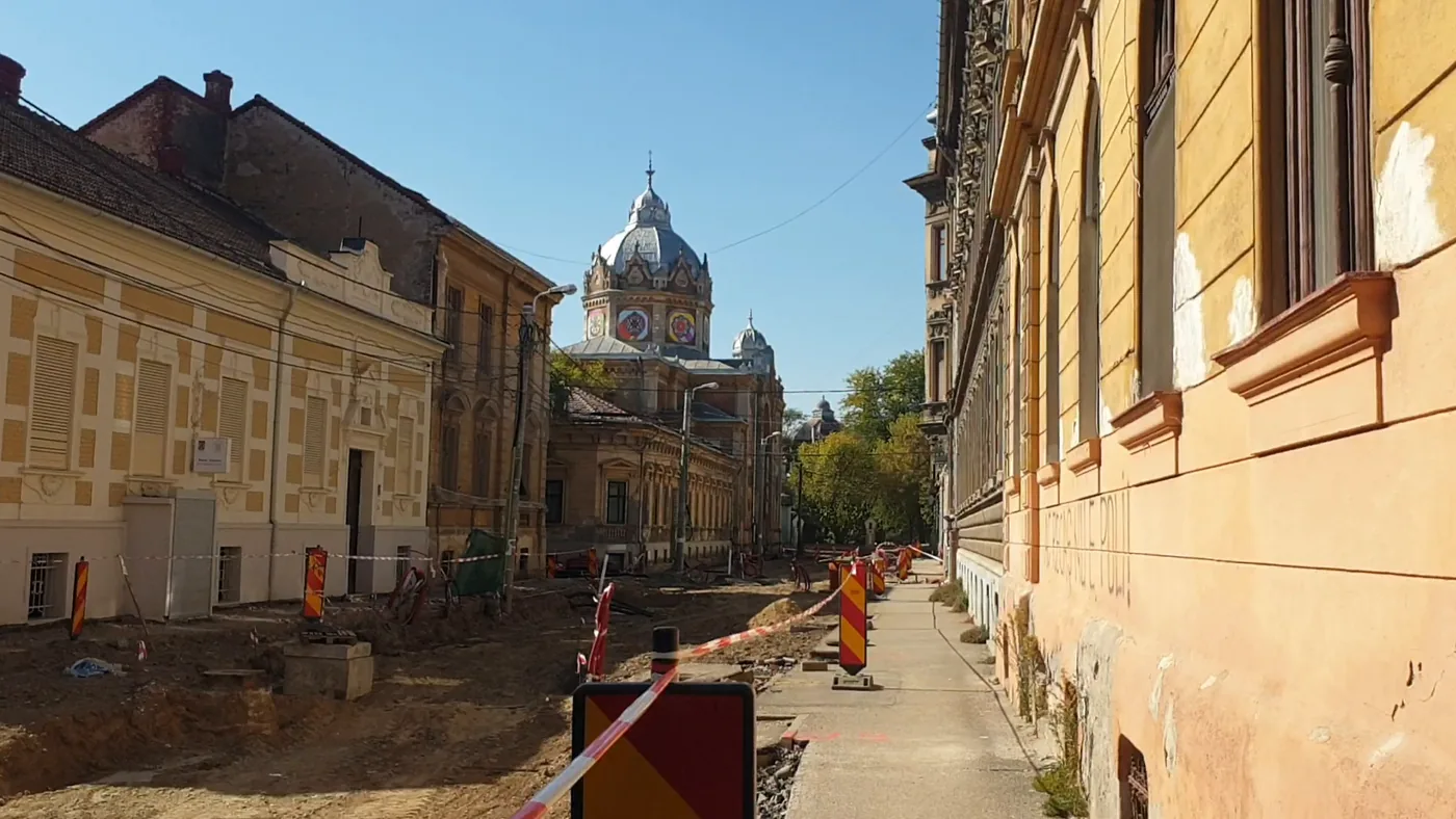 A narrow street with old houses, the roadway dug up behind red and white tape, a domed church in the distance