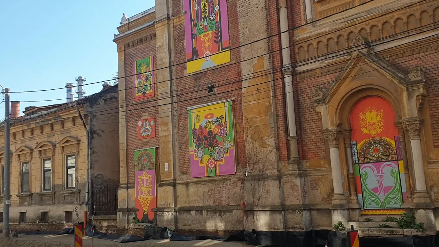 A red-brick synagogue with arched windows filled with colourful painted panels, an old single-storey house beside it