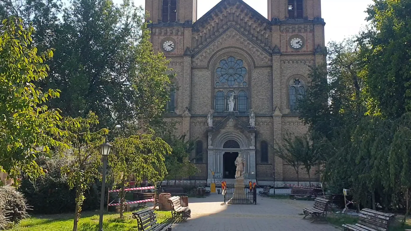 A large brick church with two tall towers and clock faces, a rose window over the entrance, trees and benches in front