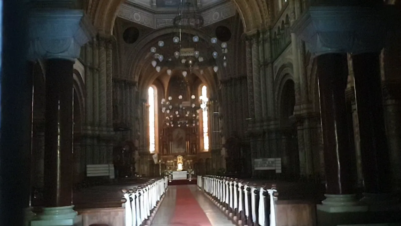 The nave of a large church seen from the entrance, dark columns, rows of pews with white covers and a red carpet to the altar