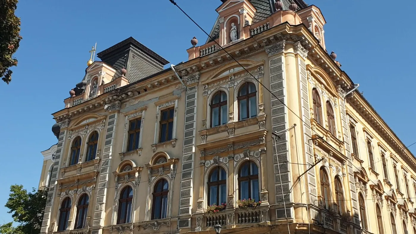 The upper floors of an ornate yellow corner building with a decorated gable, balconies and a dark mansard roof against a blue sky