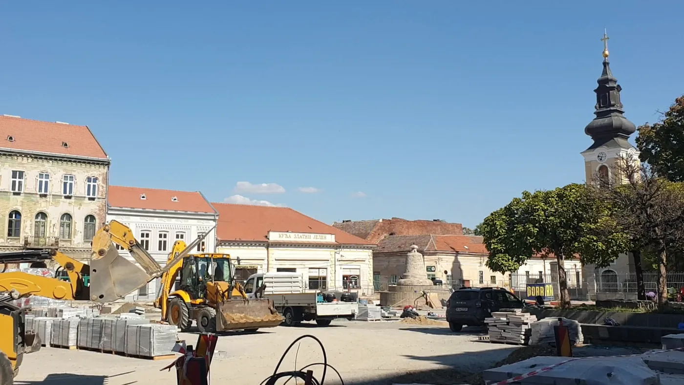 A square under reconstruction with a yellow excavator and pallets of paving stones, baroque houses and a church tower with an onion dome behind