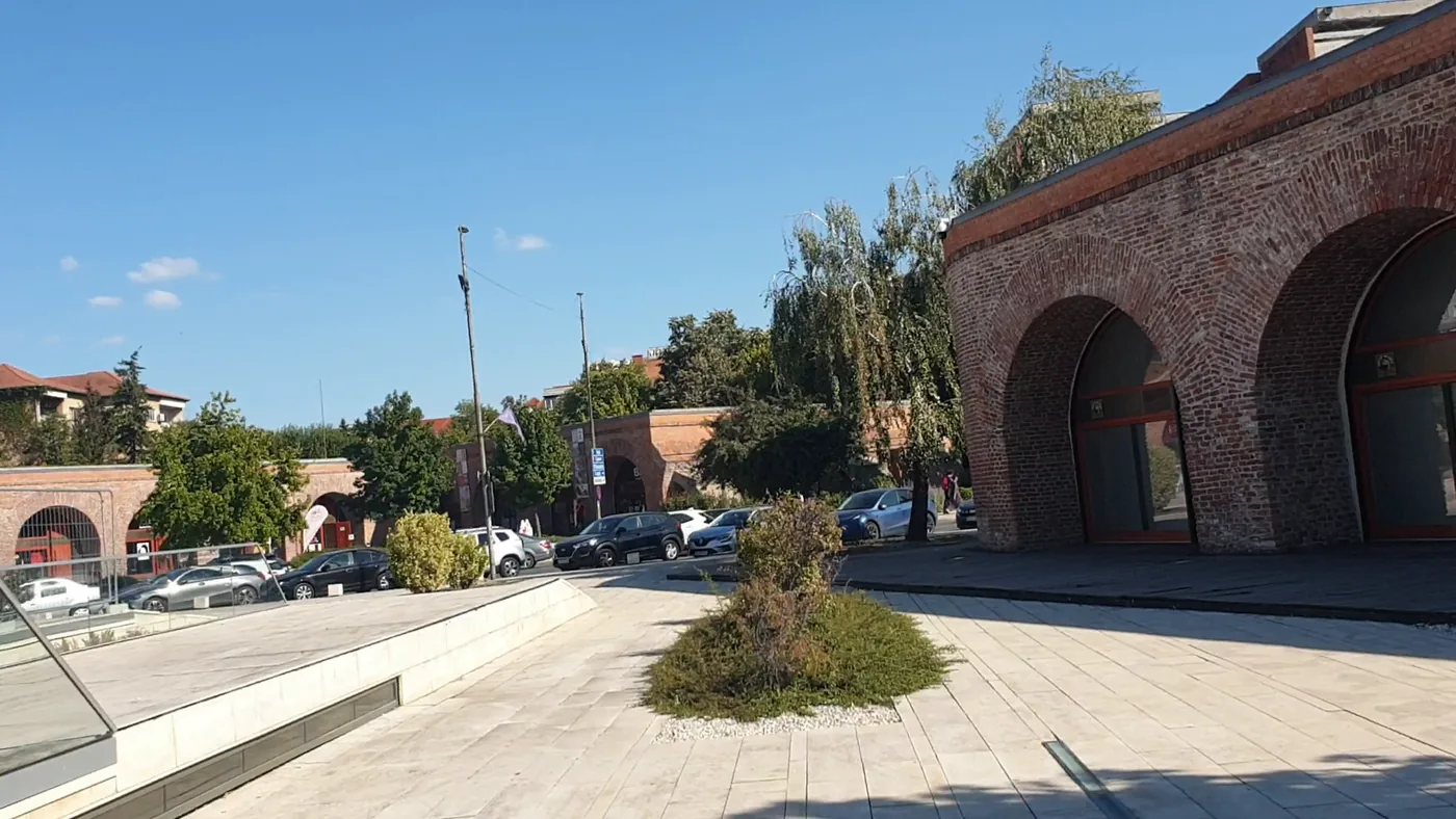 A long red-brick fortress wall with arched openings and glass doors, a paved square in front, trees and parked cars beyond