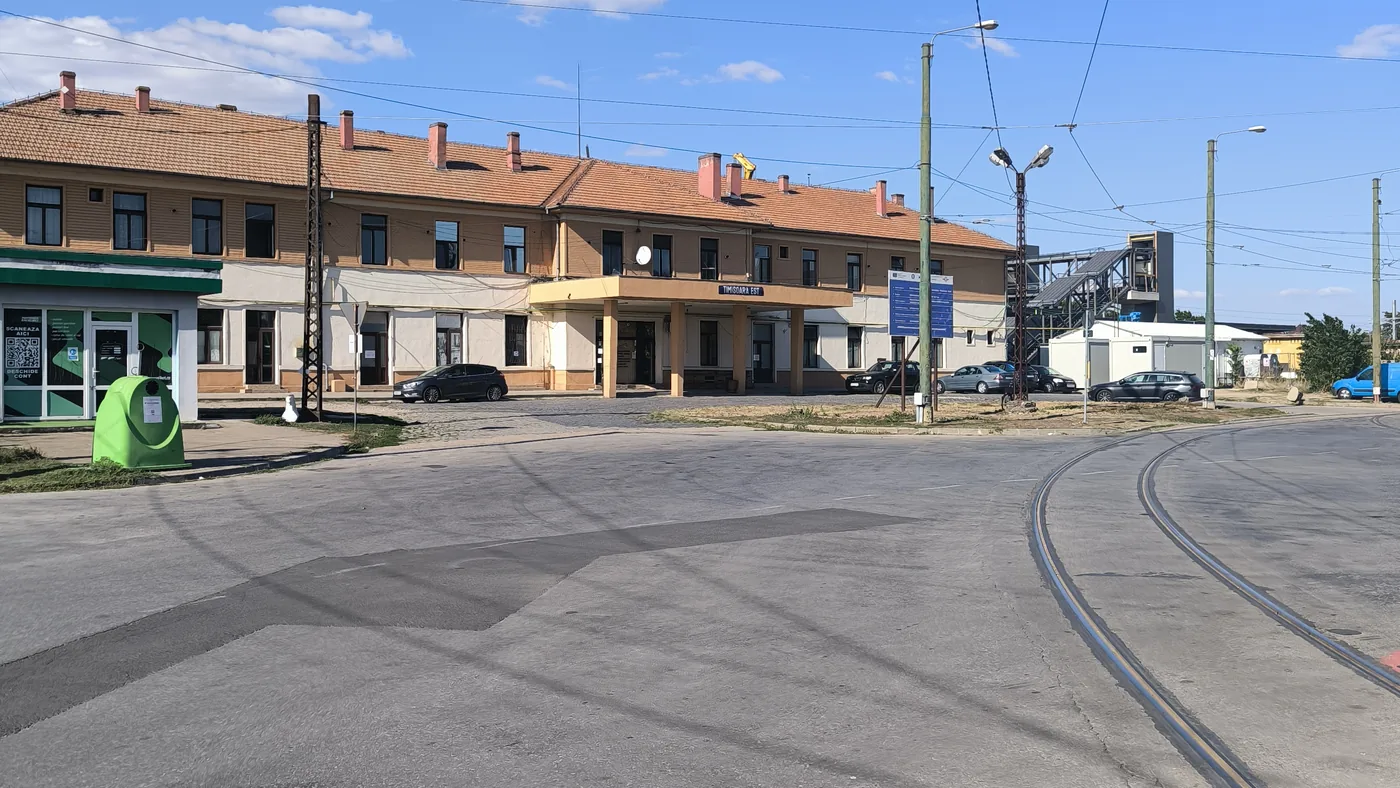 A long two-storey station building with an orange tiled roof and the sign Timisoara Est, tram tracks curving across the square in front