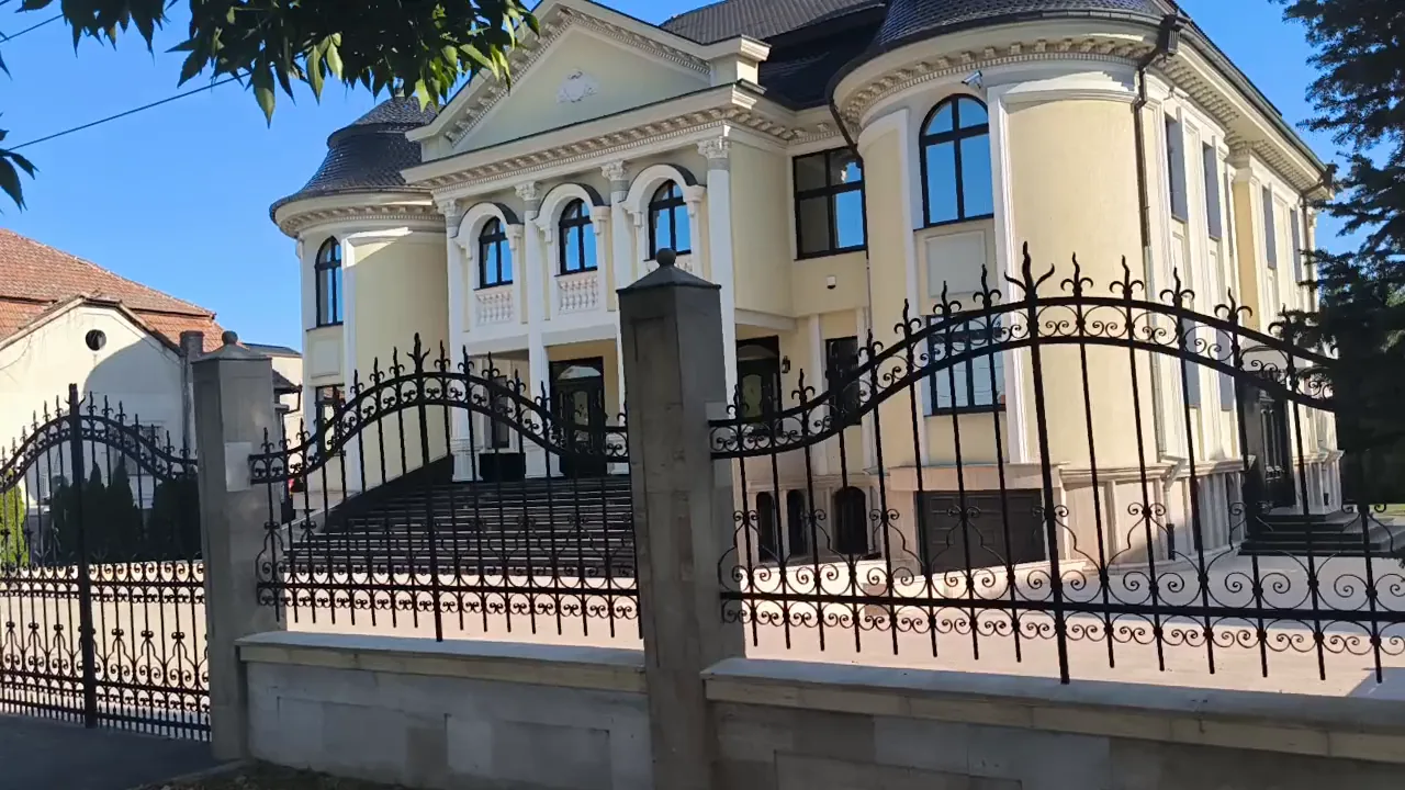 A cream-coloured villa with round corner towers and arched windows behind a black wrought-iron fence and gate