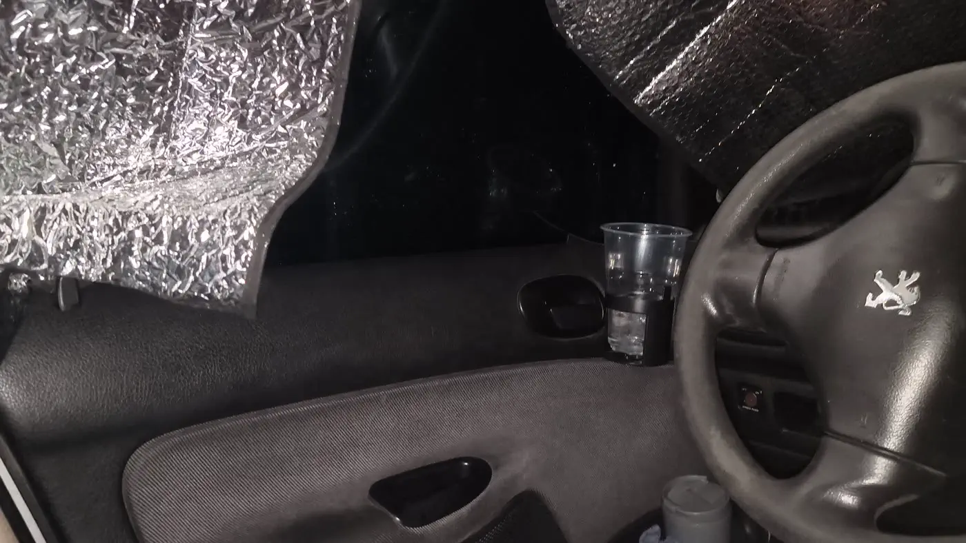 Inside a small car set up for sleeping: reflective bubble-foil sunshades covering the windows, a cup in the holder